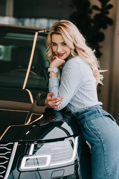 Cheerful blonde girl standing near black automobile in car showroom — Stock Photo
