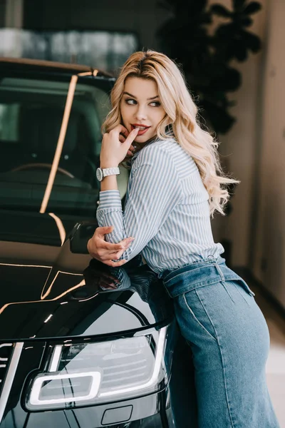 Beautiful blonde girl standing near black automobile in car showroom — Stock Photo