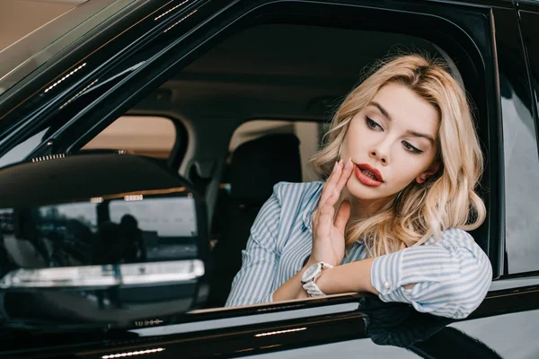 Jolie fille blonde regardant miroir de voiture tout en touchant le visage — Photo de stock