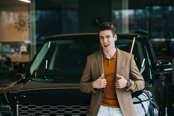 Cheerful man standing near automobile in car showroom and showing thumbs up — Stock Photo