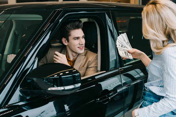 Cheerful man looking at blonde girl holding dollar banknotes in car showroom — Stock Photo