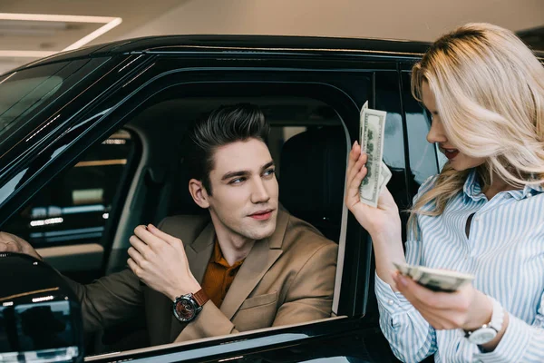 Handsome man looking at blonde girl holding dollar banknotes in car showroom — Stock Photo