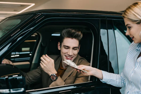 Happy man looking at dollar banknotes in hands of blonde girl in car showroom — Stock Photo