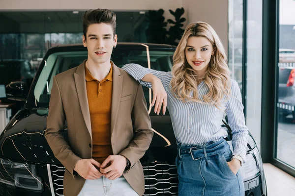 Hombre guapo y alegre chica rubia de pie con la mano en el bolsillo cerca del coche - foto de stock