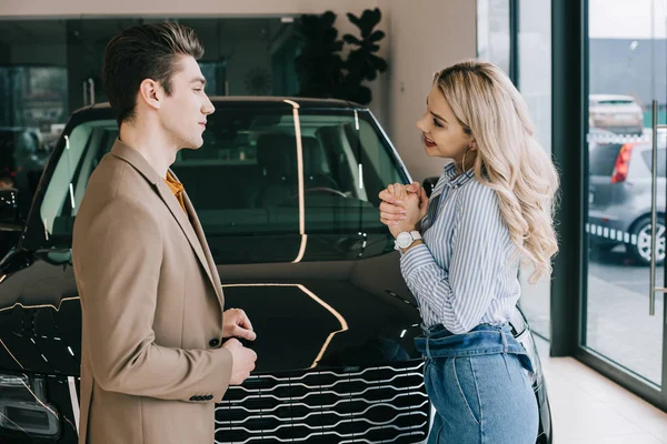 Bel homme regardant gai fille blonde tout en se tenant avec les mains serrées près de la voiture — Photo de stock