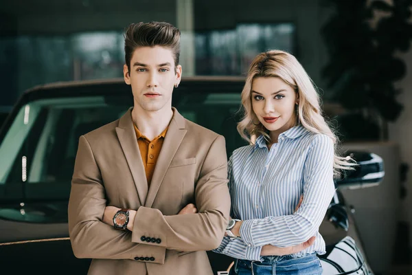 Handsome man and beautiful blonde woman standing with crossed arms in car showroom — Stock Photo