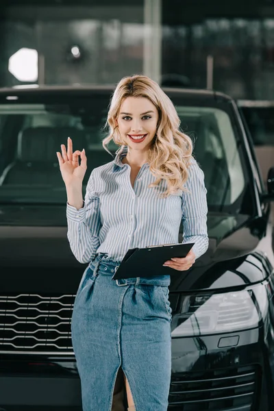 Fröhliche blonde Autohändler hält Klemmbrett und zeigt ok Zeichen in Auto-Showroom — Stockfoto