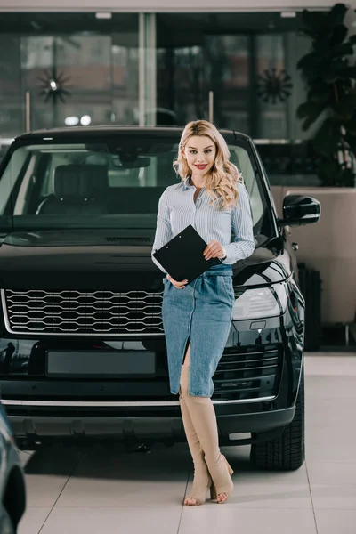 Happy blonde car dealer holding clipboard while standing in car showroom — Stock Photo