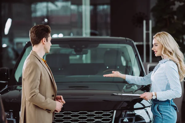 Cheerful blonde car dealer showing automobile to handsome man in car showroom — Stock Photo