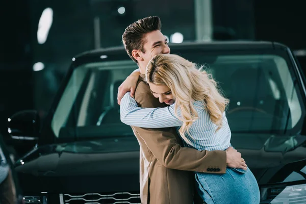 Веселая блондинка обнимает счастливого мужчину, стоя возле машины — стоковое фото