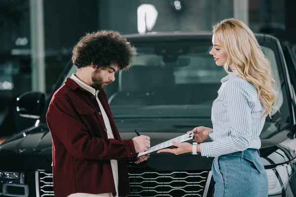 Blonder Autohändler hält Klemmbrett, während lockiger Mann Vertrag unterschreibt — Stockfoto