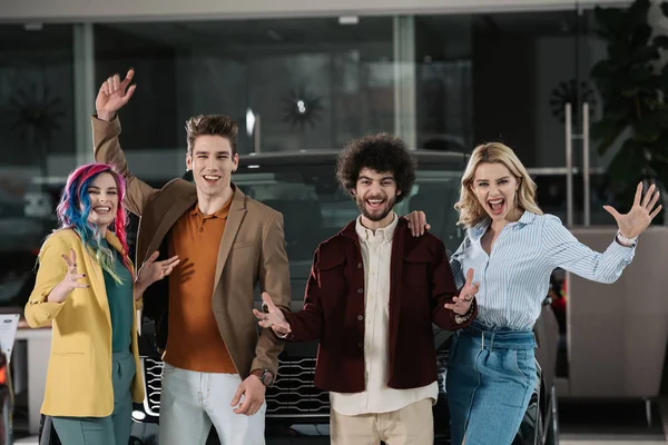 Happy group of friends gesturing near automobile in car showroom — Stock Photo