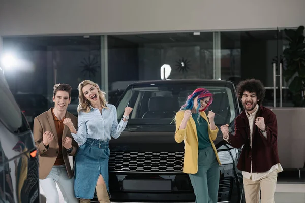 Cheerful group of friends gesturing near automobile in car showroom — Stock Photo