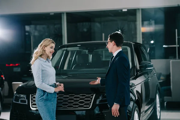 Negociante de carro mostrando automóvel para mulher loira alegre no showroom do carro — Fotografia de Stock