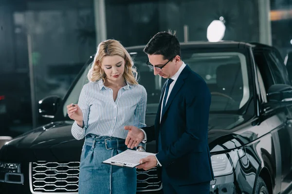 Concesionario de coches guapo sujetando portapapeles cerca atractiva mujer rubia con pluma - foto de stock