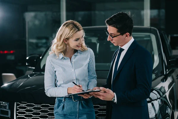 Cheerful blonde woman signing contract and looking at car dealer — Stock Photo
