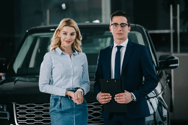 Schöner Autohändler mit Brille und Klemmbrett in der Nähe attraktiver blondes Mädchen — Stockfoto