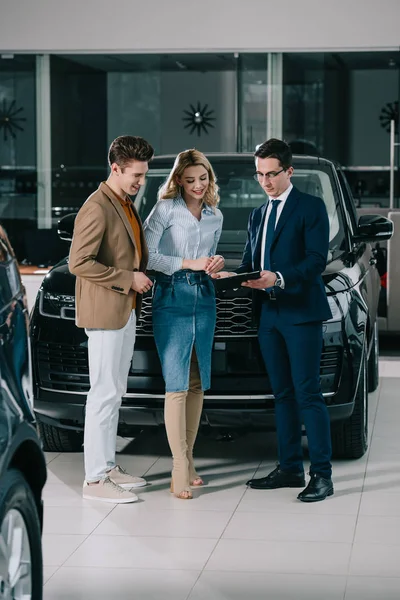 Happy man standing with blonde woman signing contract with car dealer — Stock Photo