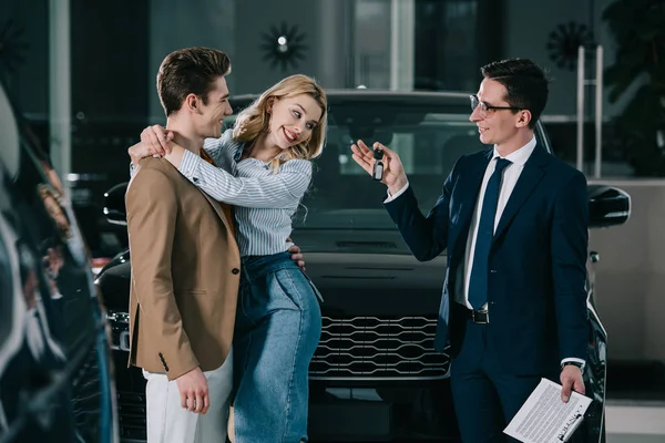 Schöner Autohändler übergibt Schlüssel an glückliche blonde Frau, die Mann umarmt — Stockfoto
