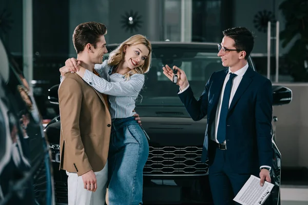 Vendedor de coches guapo dando la llave a la mujer rubia feliz abrazando al hombre - foto de stock