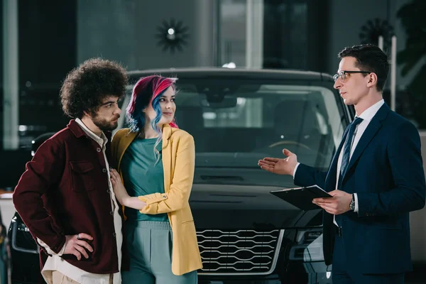 Schöner Autohändler mit Brille, Klemmbrett in der Hand und gestikulierend in der Nähe von Mann und Frau — Stockfoto