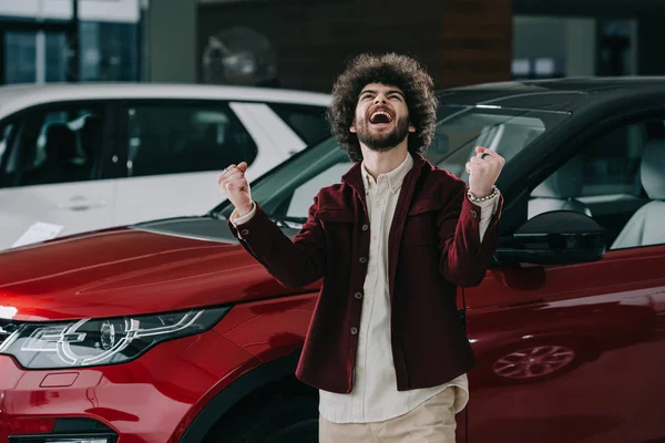 Веселый кудрявый человек жестикулирует, стоя рядом с красной машиной — стоковое фото