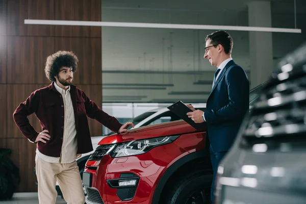 Cheerful car dealer standing near red automobile and curly man — Stock Photo