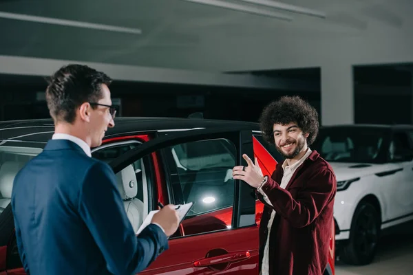 Cheerful curly man looking at car dealer in glasses while standing near red car — Stock Photo