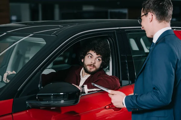 Красивий кучерявий чоловік сидить у червоній машині біля продавця автомобілів — стокове фото