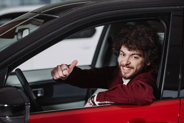 Homem encaracolado alegre sentado no carro e mostrando polegar para cima — Fotografia de Stock