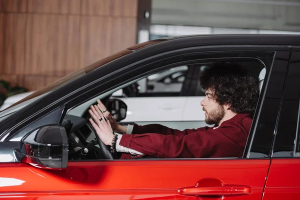 Handsome curly driver sitting in red automobile in car showroom — Stock Photo