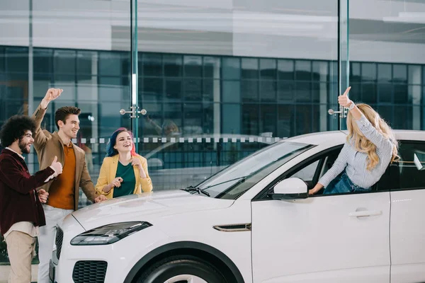 Donna bionda mostrando pollice verso l'alto vicino amici felici in auto showroom — Foto stock