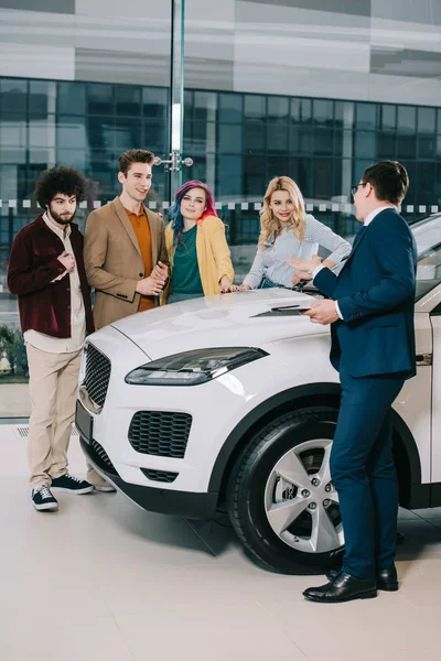 Concesionario de coches haciendo gestos mientras habla con amigos alegres cerca de coche blanco - foto de stock