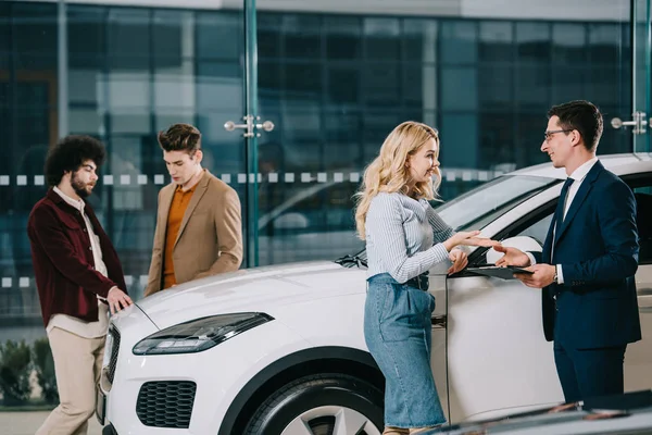 Handsome car dealer gesturing while talking with attractive blonde woman in car showroom — Stock Photo