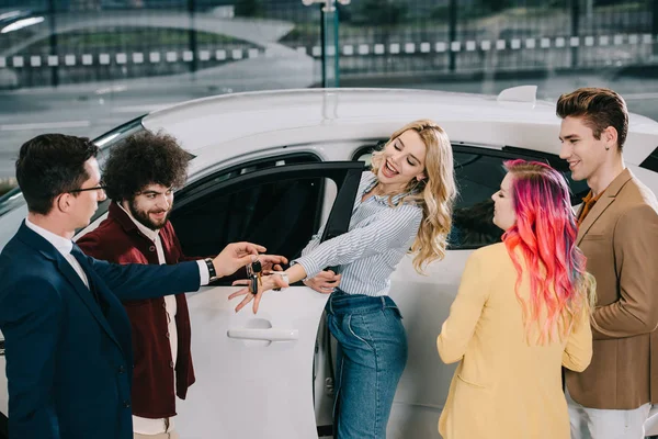 Car dealer giving key to happy blonde girl near friends in car showroom — Stock Photo