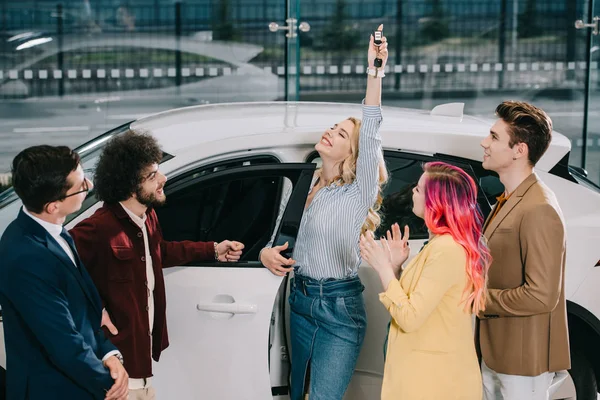 Car dealer looking at happy blonde girl holding key near friends in car showroom — Stock Photo