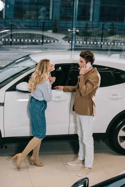 Mulher loira atraente de pé perto do homem falando om smartphone perto de carro branco — Fotografia de Stock