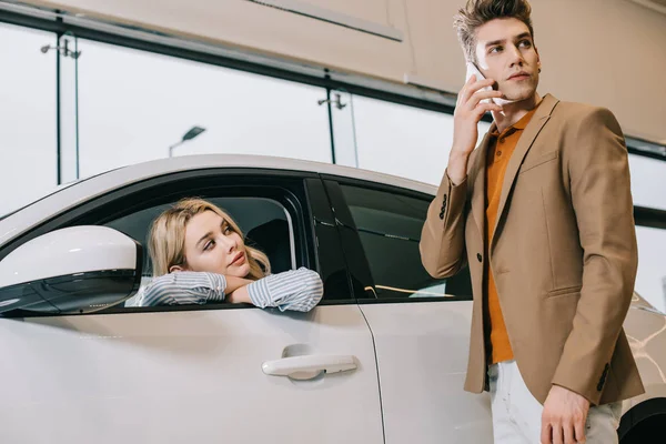 Rubia chica sentado en coche y mirando guapo hombre hablando en smartphone - foto de stock