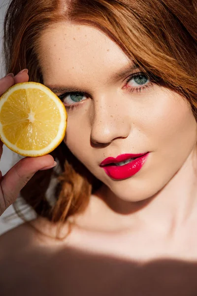 Hermosa pelirroja con labios rojos posando con limón cortado y mirando a la cámara - foto de stock