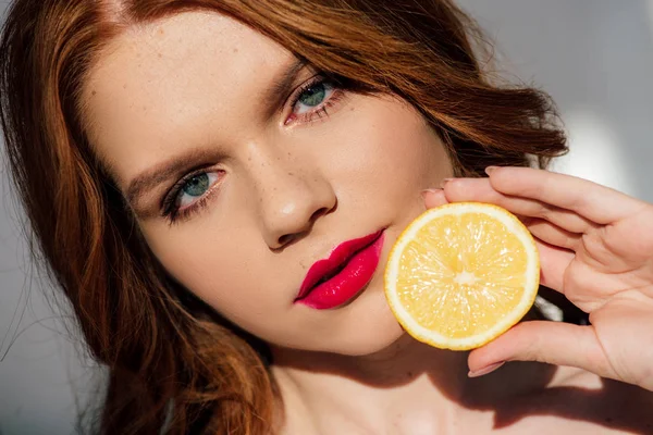 Красива руда дівчина з червоними губами дивиться на камеру і позує з лимоном — стокове фото