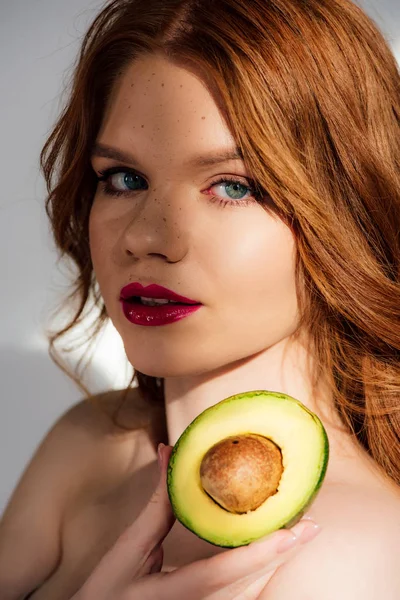 Красивая рыжая девушка с красными губами, смотрящая в камеру и позируя с вырезанным авокадо — стоковое фото