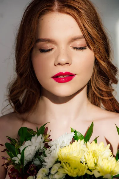 Beautiful young redhead woman with red lips and eyes closed posing with flowers — Stock Photo