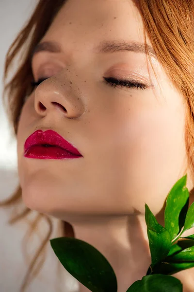 Portrait de belle jeune rousse aux lèvres rouges et aux yeux fermés posant avec une feuille verte — Photo de stock
