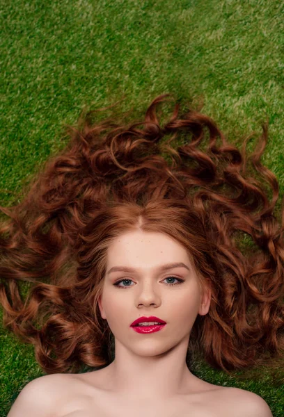 Draufsicht der schönen jungen rothaarigen Frau mit roten Lippen auf grünem Gras, die in die Kamera schaut — Stockfoto