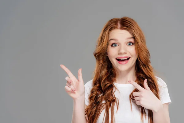 Surprised redhead girl looking at camera and pointing with fingers isolated on grey with copy space — Stock Photo