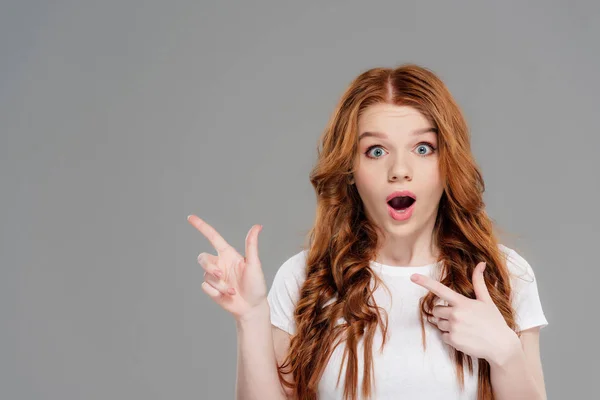Surprised redhead girl looking at camera and pointing with fingers isolated on grey with copy space — Stock Photo