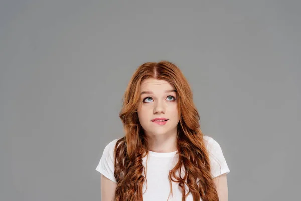 Beautiful confused redhead girl biting lip and looking up isolated on grey with copy space — Stock Photo