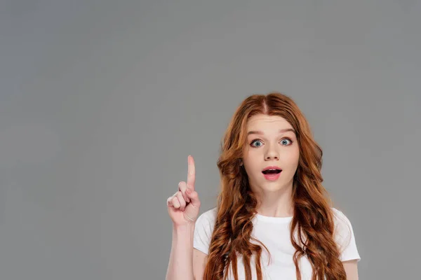 Beautiful surprised redhead girl looking at camera and showing idea gesture isolated on grey with copy space — Stock Photo