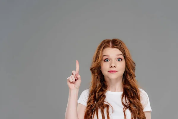 Beautiful surprised redhead girl looking at camera and showing idea gesture isolated on grey with copy space — Stock Photo