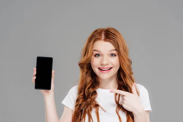 Belle rousse fille pointant du doigt le smartphone avec écran blanc, regardant la caméra et souriant isolé sur gris — Photo de stock